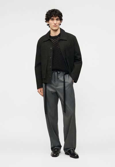 Νέος άνδρας που φοράει μαύρο σακάκι με κουμπιά, μαύρο πλεκτό πουλόβερ με υφή, γκρί παντελόνι με φαρδιές ρίγες και μακριές μαύρες κορδέλες, και μαύρα παπούτσια.