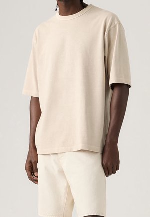 Mann trägt ein schlichtes beiges oversized T-Shirt mit kurzen Ärmeln und helle Shorts vor einem neutralen Hintergrund.