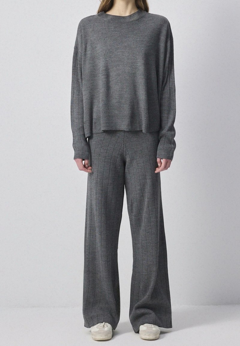 Femme portant un pull en tricot gris foncé ample et un pantalon large assorti, avec des baskets blanches, debout devant un fond clair uni.