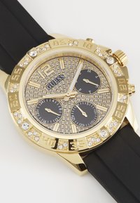 Reloj de tono dorado con acentos de rhinestones, correa de goma negra, esfera texturizada con subesferas doradas y negras, números audaces y características de cronógrafo.