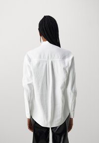 White long-sleeve shirt with a relaxed fit, featuring a back yoke seam and a curved hem, paired with black textured pants.
