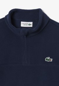 Pullover in fleece di un blu scuro, con collo alto, zip a metà sulla parte anteriore e logo Lacoste ricamato sul petto. Texture morbida con una finitura liscia.