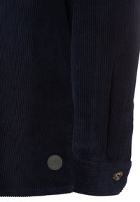 Marineblauwe ribfluwelen shirt met brede ribstructuur, voorzien van een knoopmanchet en een cirkelvormige logo patch aan de onderkant.