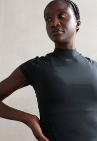 Femme aux cheveux tressés portant un haut noir ajusté, regardant sur le côté, une main reposant sur son bas du dos.