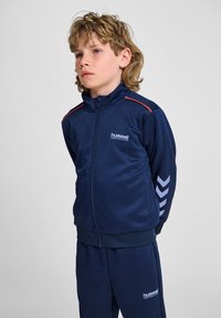 Marineblaue Trainingsjacke mit roten Akzenten, hohem Kragen, Reißverschluss vorne und Logo. Verfügt über Kettenmuster an den Ärmeln und passende Hosen.