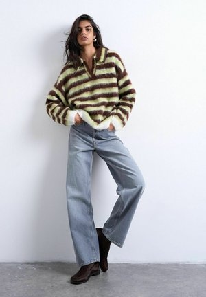 Vrouw staand met handen in haar zakken, gekleed in een bruine en lichtgroene gestreepte, donzige trui, wijde blauwe jeans en donkerbruine laarzen.
