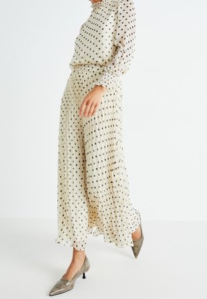 Femme portant un chemisier crème à pois à manches longues et une jupe maxi plissée assortie avec des talons bas métalliques à bout pointu.