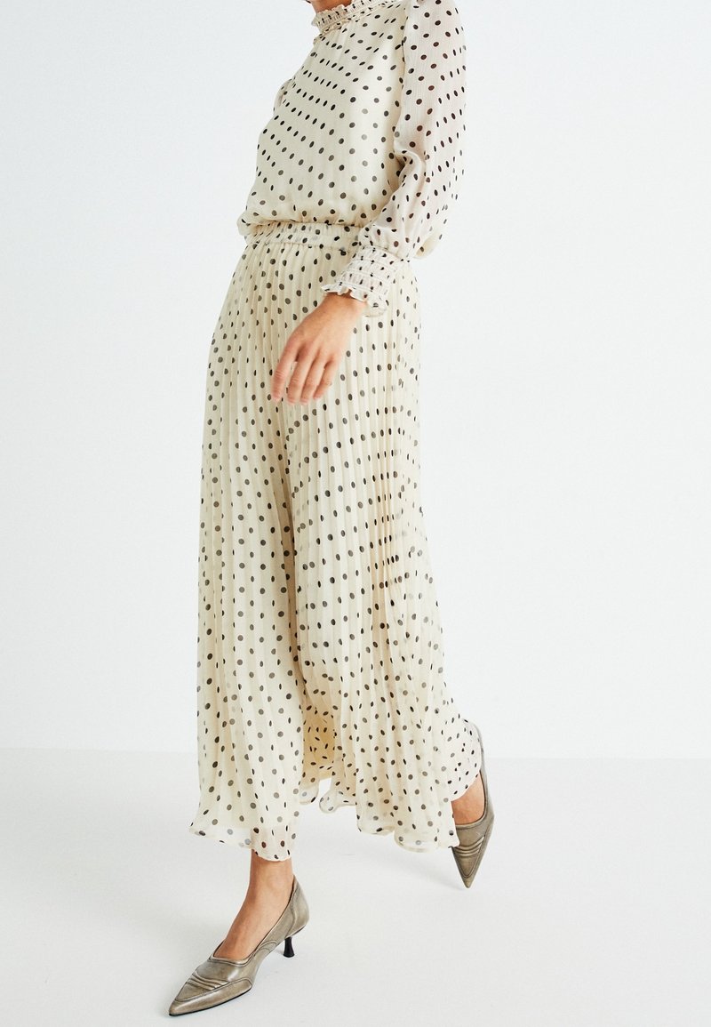 Femme portant un chemisier crème à pois à manches longues et une jupe maxi plissée assortie avec des talons bas métalliques à bout pointu.