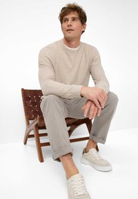 Beiger Strickpullover, hellgraue Hose und cremefarbene Sneaker. Die Sitzgelegenheit ist ein Holzstuhl mit einem geflochtenen Ledersitz. Schlichte Designelemente.