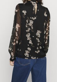 Blouse fleurie noire en tissu léger et transparent avec manches longues, motif floral en crème et rose, et détail dos avec ouverture en forme de goutte.