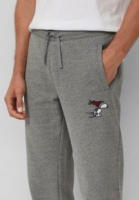 Man in grijze joggingbroek met een Snoopy-graphic met een rode pet op de dij, handen ontspannen met één hand in de zak tegen een effen achtergrond.