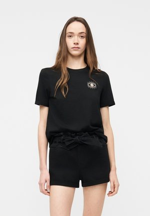 Fiatal nő hosszú barna hajjal, fekete pólót visel, amelyen hímzett szem minta látható, és fekete, magas derekú rövidnadrágot, amelyet kötött öv díszít.