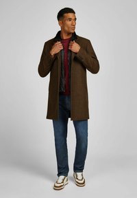 Brauner Wollmischungs-Mantel mit dezentem Muster, getragen über einer schwarzen Daunenweste. Kombiniert mit blauen Jeans und Sneakers mit braunen Akzenten.
