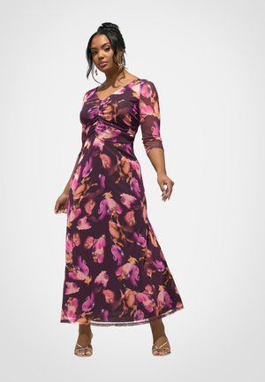 Vrouw staand, gekleed in een lange jurk met bloemenpatroon in paars, roze en oranje, driekwart mouwen en gouden sandalen met bandjes.