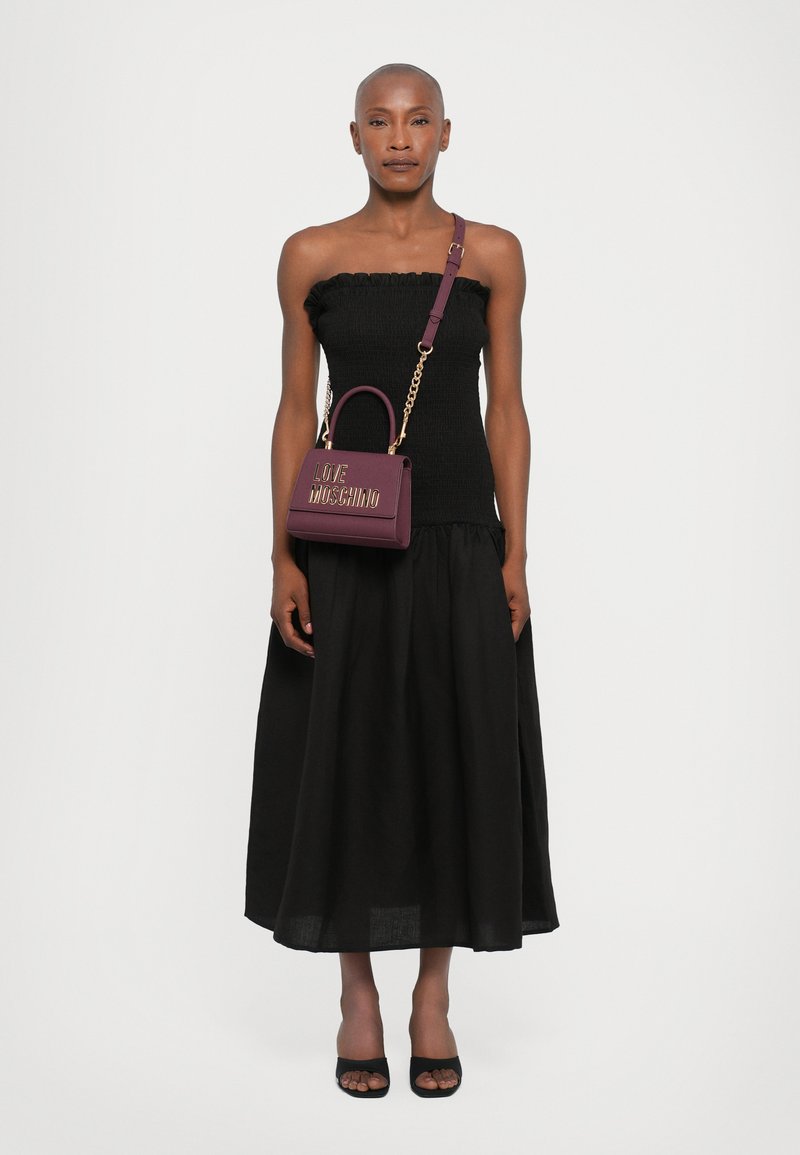 Zwarte strapless top met ruffle detail, gecombineerd met een zwarte rok. Houdt een bordeaux leren handtas met gouden opdruk vast, crossbody gedragen.