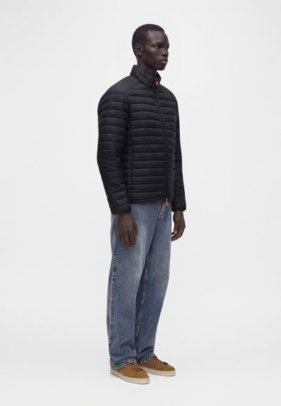 Homme portant une veste matelassée noire, un jean bleu ample et des baskets marron, debout devant un fond blanc uni.