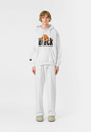 Sudadera blanca con estampado gráfico y acentos naranjas, combinada con pantalones blancos holgados y zapatos de textura verde claro. Conjunto casual.