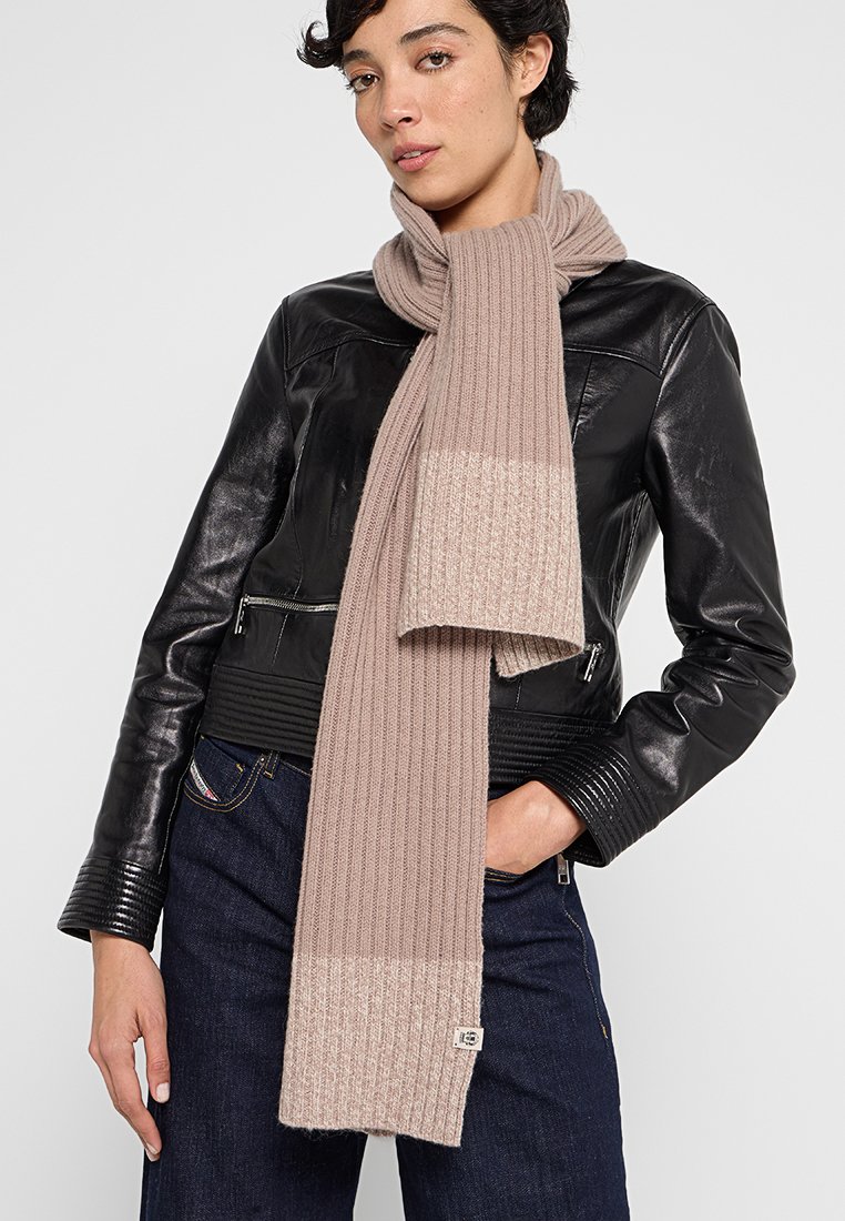 Beżowy sweter w formie szalika, z prążkowaną fakturą i jednolitym jaśniejszym dołem. Noszony z czarną skórzaną kurtką, wyeksponowany na neutralnym tle.