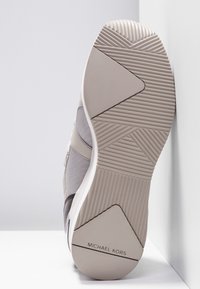 Semelle d'une basket beige et blanche avec motifs de grip texturés et logo "Michael Kors" sur le talon, placée verticalement contre un mur.