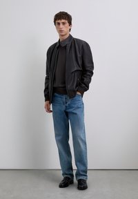 Junger Mann in einer schwarzen Lederjacke, dunklem Sweater, blauen Jeans und schwarzen Schuhen, der vor einer schlichten weißen Wand steht.