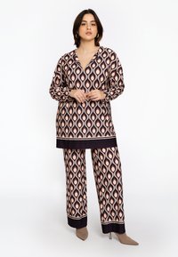Vrouw in een bijpassende tuniek en wijde broek met zwart en beige druppelvormen, staande tegen een witte achtergrond.