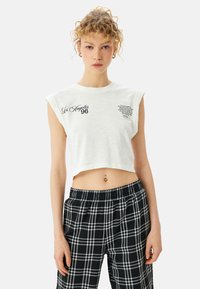 Camiseta blanca corta con cuello redondo, mangas cortas y texto gráfico "Los Ángeles 96" en la parte delantera. Combinada con pantalones de cuadros en blanco y negro.