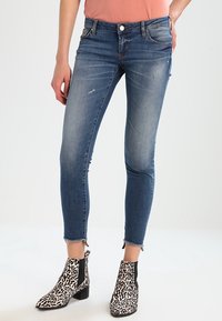 Jeans de denim azul ajustados con detalles desgastados, una cintura a media altura y dobladillos deshilachados, combinados con botas de tobillo de estampado de leopardo en blanco y negro.