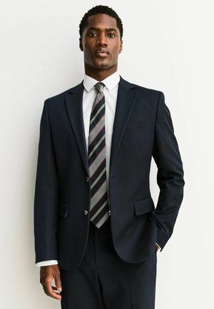 Bărbat purtând un costum bleumarin închis, cu o cămașă albă și o cravată cu dungi, stând cu o mână în buzunar, pe un fundal alb simplu.