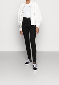 Chaqueta bomber blanca sobre una camiseta ajustada blanca, combinada con jeans negros ajustados de tiro alto y zapatillas negras y blancas. Texturas suaves.