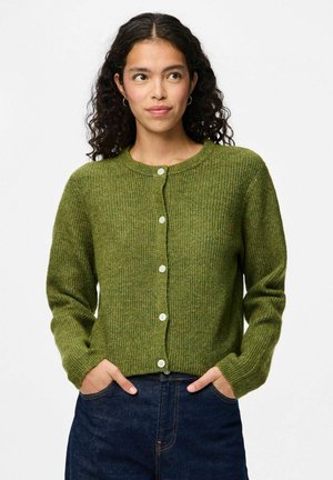 Cardigan en tricot vert avec un col rond, texture côtelée, fermée par des boutons à l'avant et des manches longues. Porté sur un denim bleu, mains dans les poches.