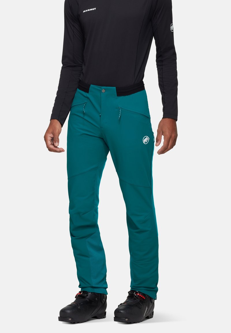Türkise Outdoor-Hose mit glatter Textur, Reißverschluss-vordertaschen und Logodetails. Kombiniert mit einem schwarzen Langarmoberteil und schwarzen Stiefeln.