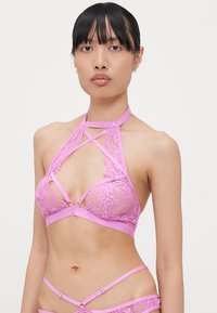 Pink lace bralette with a halter neck design, featuring crisscross straps and satin accents. Lace has a floral pattern and a soft texture.