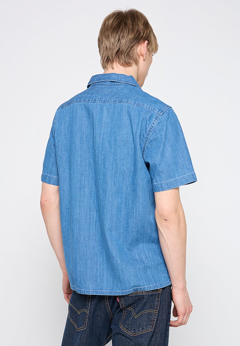 Jeune homme de dos portant une chemise en jean bleu clair à manches courtes et un jean bleu foncé devant un fond blanc uni.