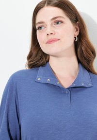 Sininen polo-paita nappikauluksella, jossa on metalliklipsejä ja teksturoitua kangasta. Suunnittelu on rento ja epämuodollinen.