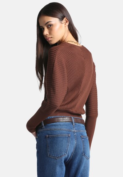 Jeune femme regardant par-dessus son épaule, portant un pull côtelé marron, un jean bleu avec une ceinture marron, et un collier chaîne en or.