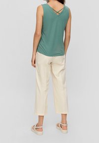 Top senza maniche verde chiaro con dettaglio incrociato nella parte posteriore, abbinato a pantaloni ampi color crema e sandali beige, con una texture liscia.