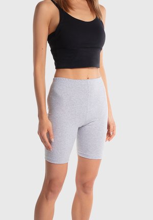 Kobieta w czarnym, krótkim topie na ramiączkach i dopasowanych, jasnoszarych szortach do kolan, stojąca na tle jasnego, gładkiego tła.