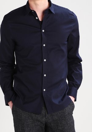 Mężczyzna w ciemnogranatowej koszuli na guziki z białymi guzikami i szarych, teksturowanych spodniach, ręce w kieszeniach, na jednolitym tle.