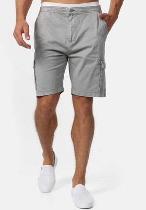 Mann trägt hellgraue Cargo-Shorts und weiße Slipper, läuft mit einer Hand in der Tasche vor weißem Hintergrund.