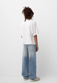 Bílé oversized tričko s krátkým rukávem, v kombinaci s světle modrými širokými džínami se sepraným vzhledem. Outfit doplňují béžové tenisky s gumovou podrážkou.