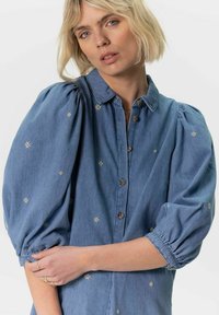 Robe en denim avec des manches bouffantes, fermeture à boutons sur le devant, et broderies florales en blanc et jaune. Texture douce et couleur bleu clair.