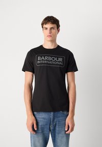 Tricou din bumbac negru cu decolteu rotund; prezintă un logo dreptunghiular gri „BARBOUR INTERNATIONAL” pe piept. Purtați cu blugi albaștri.
