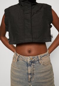 Svart kortärmad väst med huva, texturerat tyg och rynkade detaljer vid axlarna. Kombineras med högmidjade ljusa jeans.