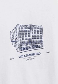 Camiseta blanca con un dibujo en línea azul marino de un edificio, texto "WILLIAMSBURG," "ESTD. 1990," y "Jack & Jones" debajo.