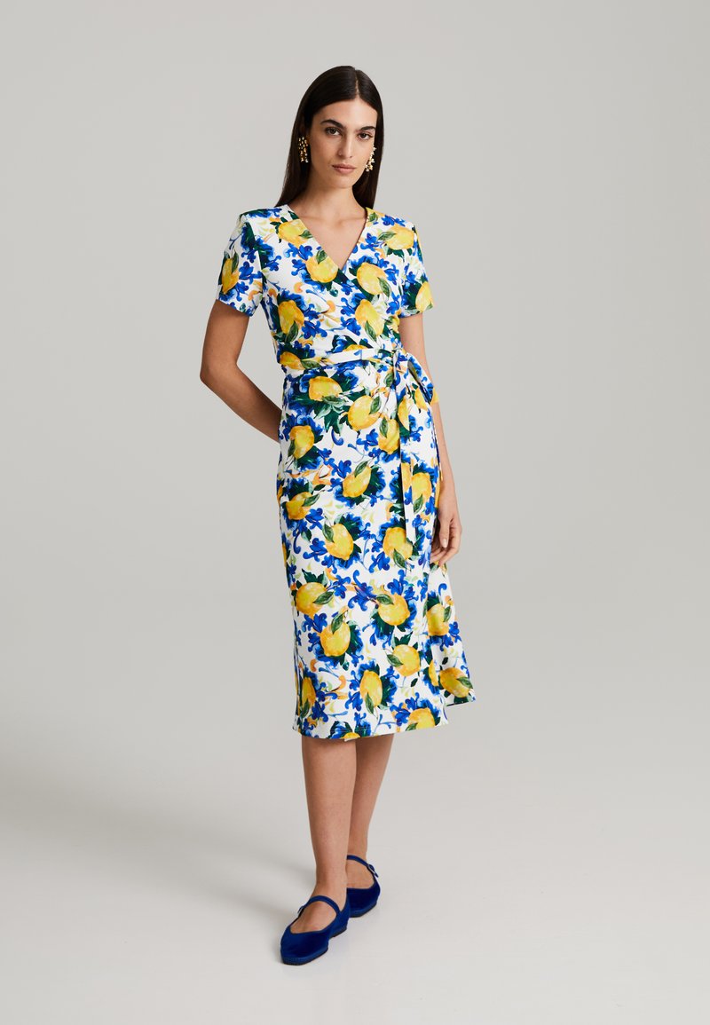 Blumenkleid mit weißem Grund, gelben Zitronen und blauen Blattmustern. V-Ausschnitt, kurze Ärmel und eine Taillenschnürung.