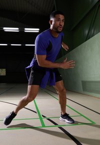 Idrettsutøver i blå t-skjorte med korte ermer og svarte shorts, som utfører en lateralt bevegelse på en bane med grønne og svarte linjer, iført svarte joggesko.