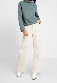 Person som bär en teal långärmad polotröja, krämfärgade vida benbyxor i kord, vita fyrkantiga stövlar, med en hand i fickan och ringar på fingrarna.