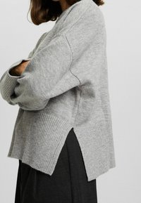 Szary dziany sweter z opadłymi ramionami, prążkowanymi mankietami i dołem, a także bocznymi rozcięciami. Miękka tekstura i luźny krój. Noszony z czarnymi spodniami.