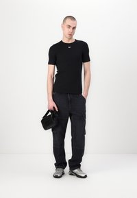Svart tettsittende t-skjorte med liten logo, mørke cargobukser med lommer, bærende en liten svart veske, og iført sølvfargede sneakers.