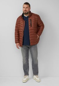 Bruine pufferjack met horizontale stiksels, hoge kraag en ritszak. Gepaard met grijze jeans en witte sneakers op een effen achtergrond.